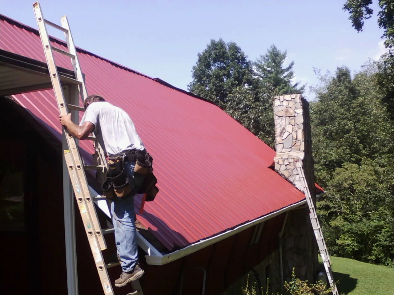 Roofer on a ladder beside a red metal roof and stone chimney — Soffit & Fascia Repair by Roofing Brothers in Carlisle